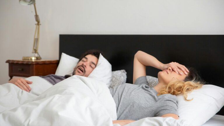 man snoring while the woman next to him covers her eyes with one hand in a sign of annoyance